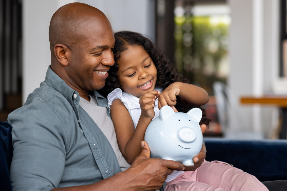 Dad holding young girl putting coins in a piggy bank