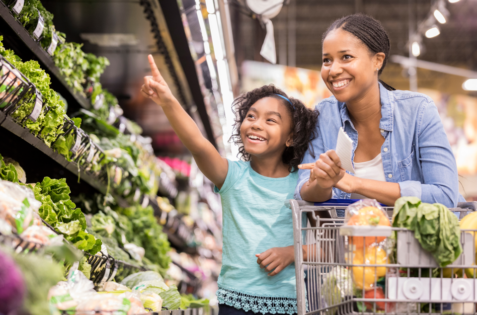 Young girl shopping with mom in produce section of grocery store.